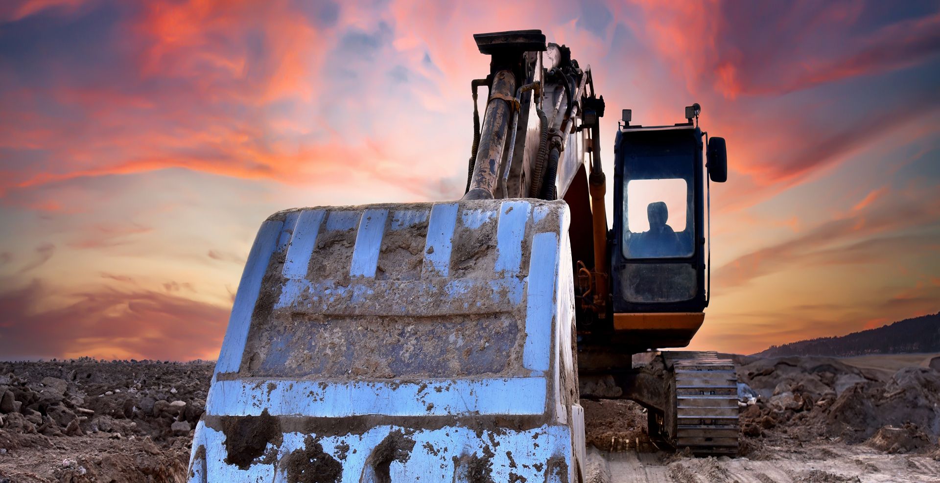 Großer Bagger mit breiter, leicht verschmutzter Baggerschaufel frontal im Abendlicht auf aufgerissener Erdfläche vor farbigem Sonnenuntergangshimmel